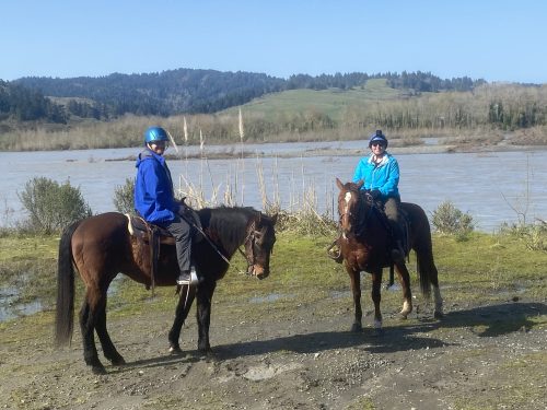 Two riders on horseback near a river, surrounded by greenery and hills under a clear blue sky.