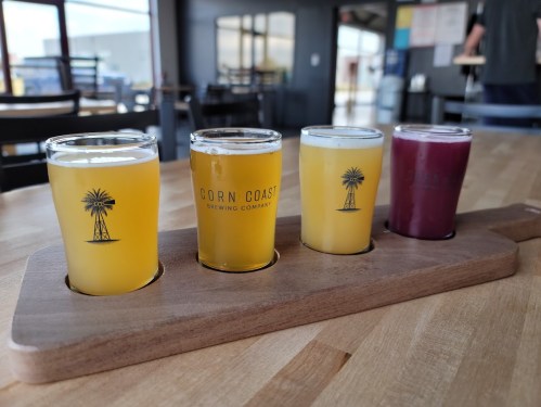 Four small glasses of beer on a wooden flight paddle, featuring different colors and styles, with a brewery logo.