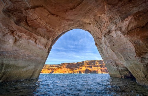 A scenic view through a rock arch, overlooking calm water and colorful cliffs under a blue sky.