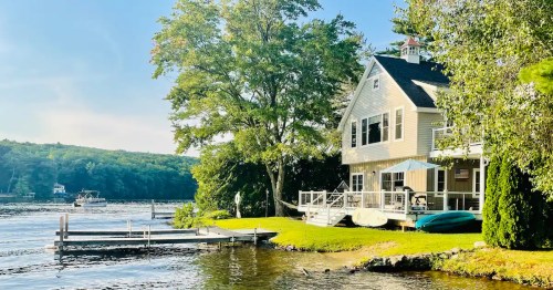 A serene lakeside view featuring a house, dock, and boats surrounded by trees under a clear blue sky.