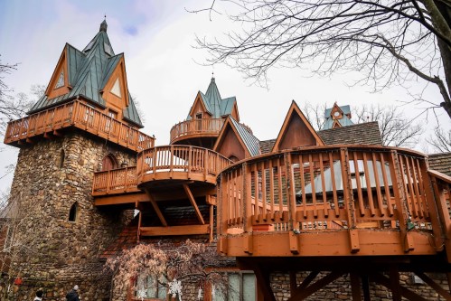 A whimsical stone building with multiple towers and wooden balconies, surrounded by bare trees against a cloudy sky.