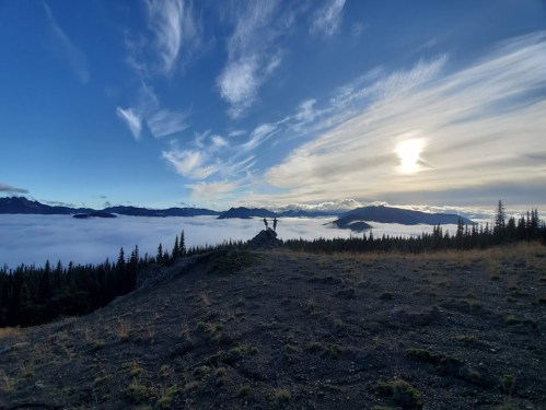 A person stands on a rocky outcrop overlooking a sea of clouds, with mountains and a sunset in the background.