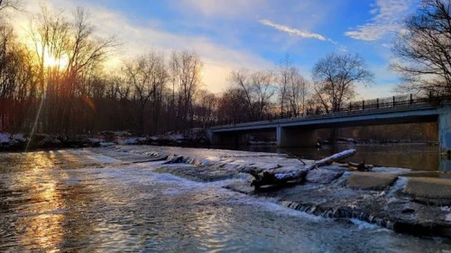 A serene river scene at sunset, with a bridge in the background and gentle water flowing over rocks.