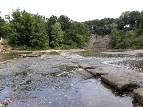 A serene river scene with rocky formations and lush green trees lining the banks under a cloudy sky.