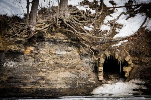 A stone archway entrance to a cave, surrounded by snow and exposed tree roots on a rocky hillside.