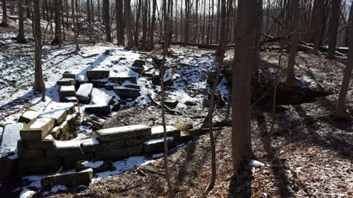 Ruins of stone structures in a snowy forest, surrounded by trees and dappled sunlight.