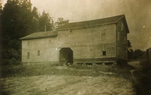 A weathered wooden building with multiple windows, set in a grassy area surrounded by trees. The image is slightly blurred.