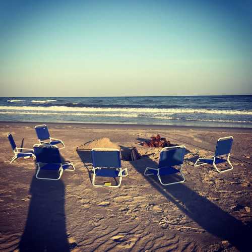 Beach scene with blue chairs arranged around a fire pit, waves gently lapping at the shore under a clear sky.