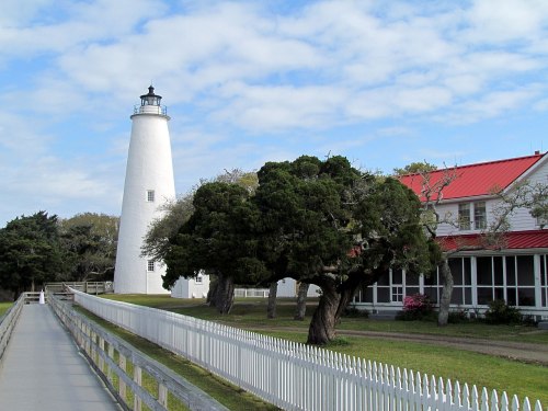 A white lighthouse beside a red-roofed building, with a path leading through green grass and trees under a cloudy sky.