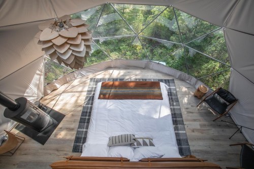 A cozy, modern geodesic dome interior with a bed, wood stove, and views of surrounding greenery.