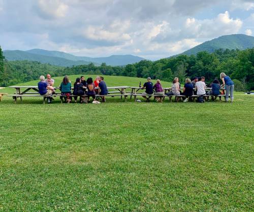 A group of people sitting at picnic tables in a grassy area, with mountains and cloudy skies in the background.