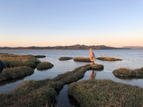 A person in a robe stands on grassy patches in a calm waterway during sunset, with hills in the background.