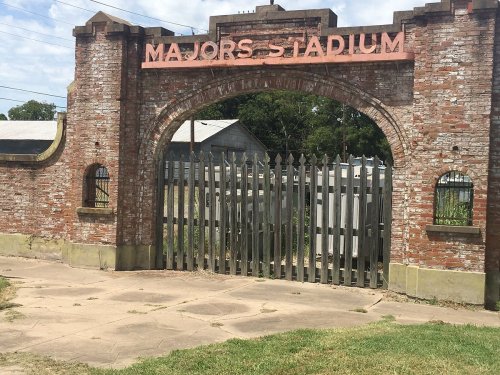 Entrance gate of Majors Stadium, featuring a brick archway and metal bars, surrounded by grassy area and trees.