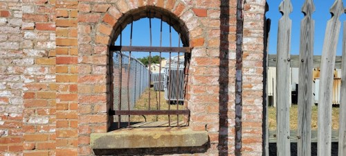 A brick wall with an arched window featuring metal bars, overlooking a fenced area and clear blue sky.