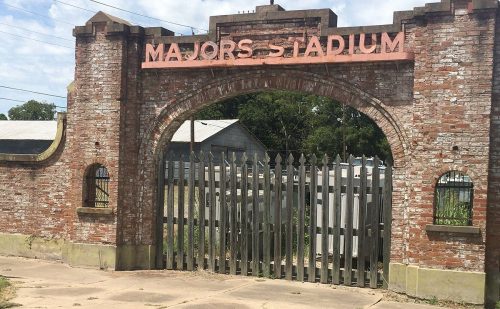 Entrance gate of Majors Stadium, featuring a brick archway and metal bars, surrounded by grassy area and trees.