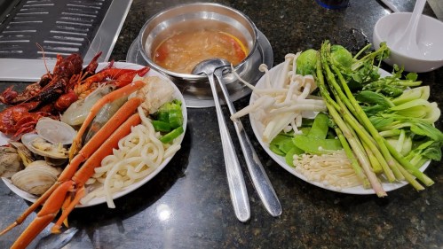 A table with two plates of fresh seafood and vegetables, alongside a pot of simmering broth.