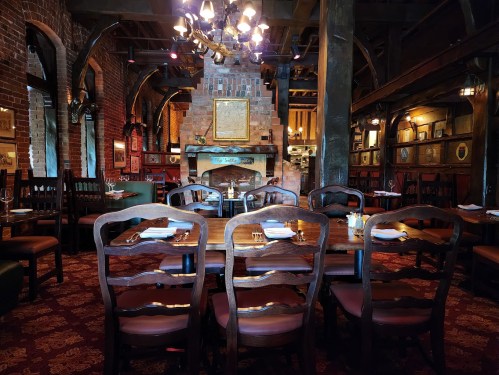 Cozy restaurant interior with wooden tables, chairs, and a brick fireplace, featuring warm lighting and rustic decor.