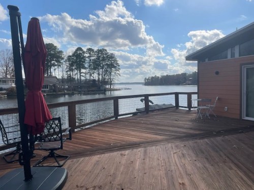A wooden deck overlooking a calm lake, with a red umbrella and trees in the background under a partly cloudy sky.