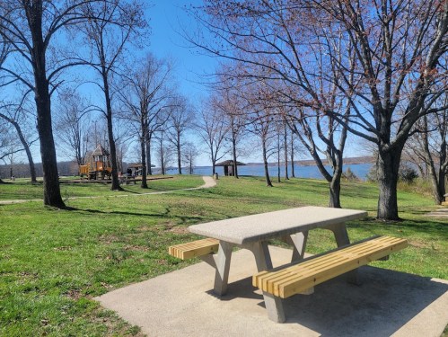 A picnic table in a park with bare trees, a path, and a lake in the background under a clear blue sky.