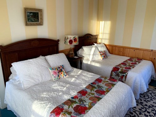 A cozy bedroom with two beds, each adorned with white bedding and colorful decorative pillows, and a floral lamp.