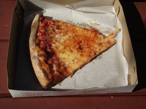 A single slice of cheese pizza in a takeout box, resting on a wooden surface.