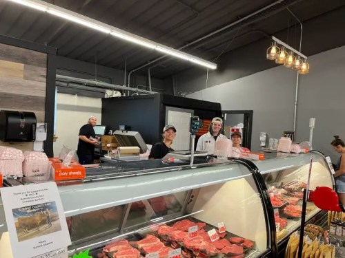 Three smiling employees stand behind a meat counter in a modern grocery store, with various cuts of meat displayed.
