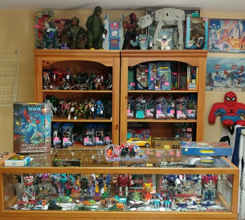 A display of vintage toys, including action figures, robots, and collectibles, in a wooden cabinet and shelves.