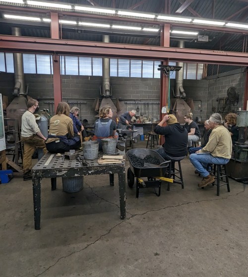 A group of people gathered in a workshop, engaged in a blacksmithing demonstration with tools and equipment around.