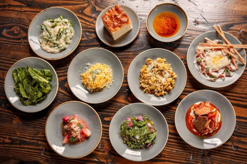 A variety of dishes arranged on plates, including pasta, greens, and a dessert, on a wooden table.
