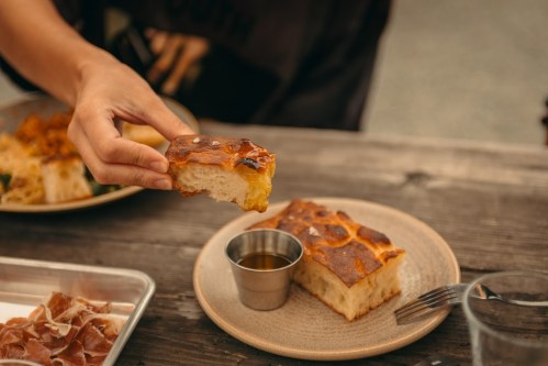 A hand holds a piece of golden-brown bread above a plate with a small cup of olive oil.