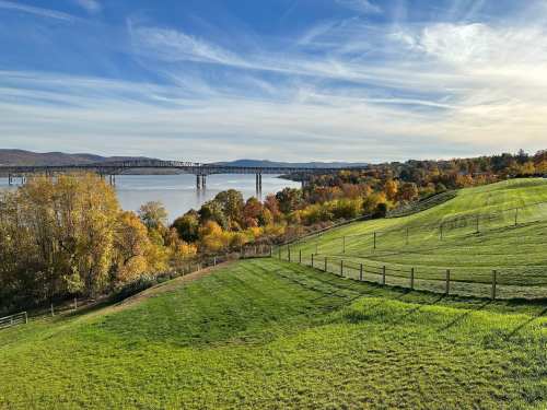 A scenic view of rolling green hills and colorful autumn trees by a river, with a bridge in the background under a blue sky.