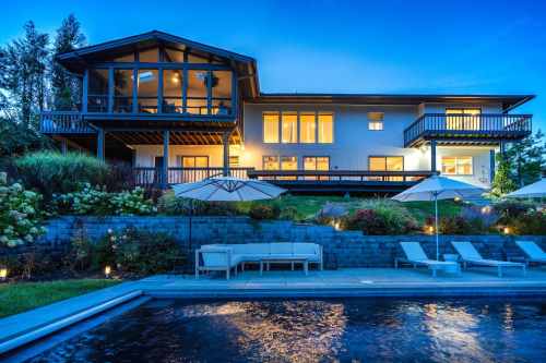 A modern house with large windows, a pool, and patio furniture, surrounded by greenery at dusk.