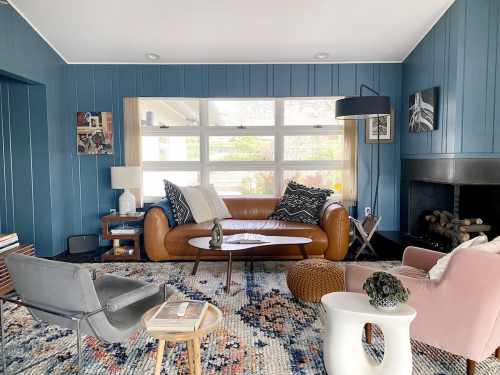 Cozy living room with blue walls, a brown leather sofa, colorful rug, and modern decor, featuring a fireplace and large windows.