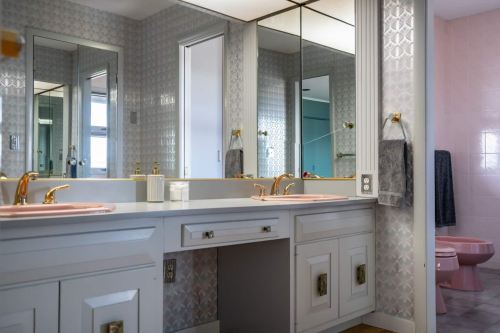 A bright bathroom featuring dual sinks, gold fixtures, and a pink toilet, with mirrored cabinets and patterned walls.