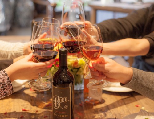 A group of hands toasting with wine glasses around a bottle of red wine on a table decorated with flowers.