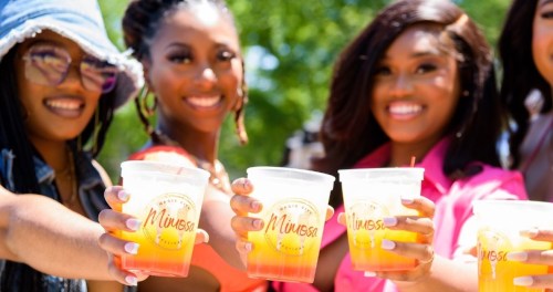 A group of friends joyfully holding colorful drinks labeled "Mimosa" in a sunny outdoor setting.