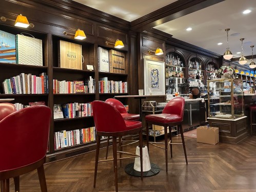 Cozy interior of a bar with wooden shelves filled with books, red chairs, and a well-stocked bar in the background.