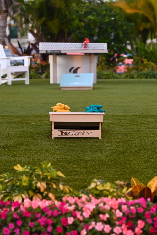 A cornhole game set up on green grass, featuring a wooden board and bags in yellow and blue, surrounded by flowers.