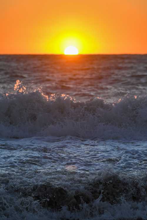 A vibrant sunset over the ocean, with waves crashing in the foreground and a warm orange sky.