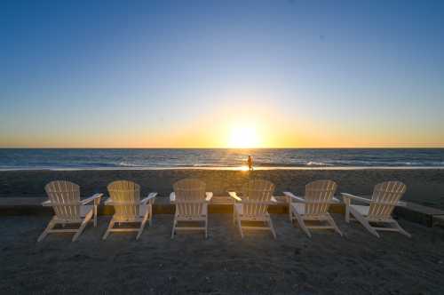 A row of white Adirondack chairs facing a sunset over the ocean, with a person walking along the shoreline.