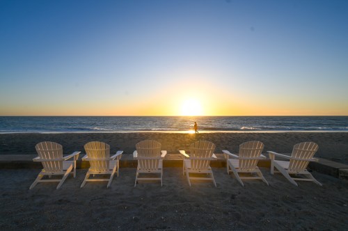 A row of white Adirondack chairs facing a sunset over the ocean, with a person walking along the shoreline.