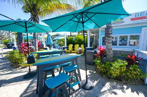 Outdoor dining area with colorful tables and chairs, shaded by teal umbrellas, surrounded by tropical plants.