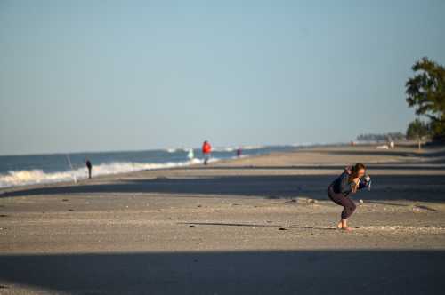 A woman crouches on a beach, taking photos, with people walking and fishing in the background along the shoreline.