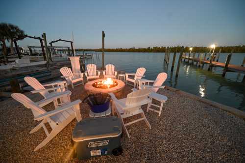 A cozy fire pit surrounded by white chairs by the water, with a dock in the background at dusk.