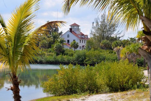 A scenic view of a house with a red roof surrounded by lush greenery and palm trees near a calm water body.