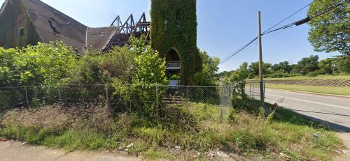 Abandoned building covered in ivy, with a damaged roof, surrounded by overgrown grass and a chain-link fence.