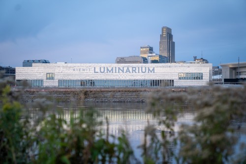 A modern building with "Kewit Luminarium" signage, reflecting on a river, surrounded by urban architecture.