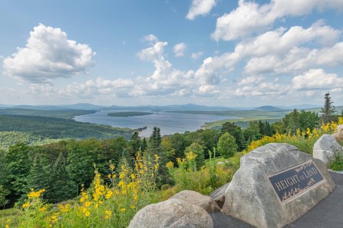 Scenic view of a lake surrounded by mountains and lush greenery, with a sign reading "Height of Land" in the foreground.