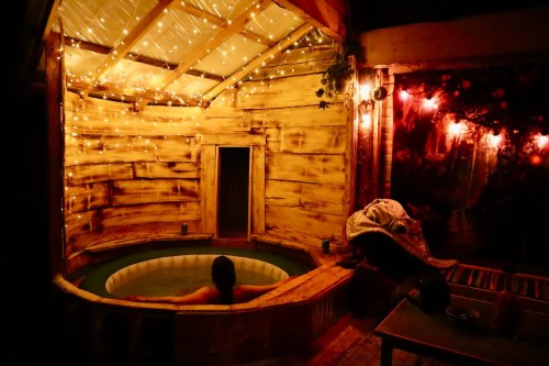 A cozy wooden hot tub surrounded by warm string lights, with a person relaxing inside.