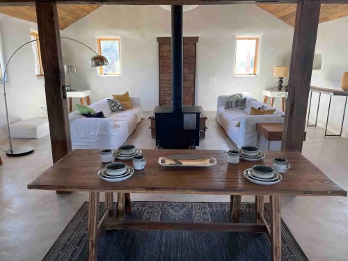 Cozy interior featuring a wooden dining table set, a wood stove, and two beds with decorative pillows in a bright space.